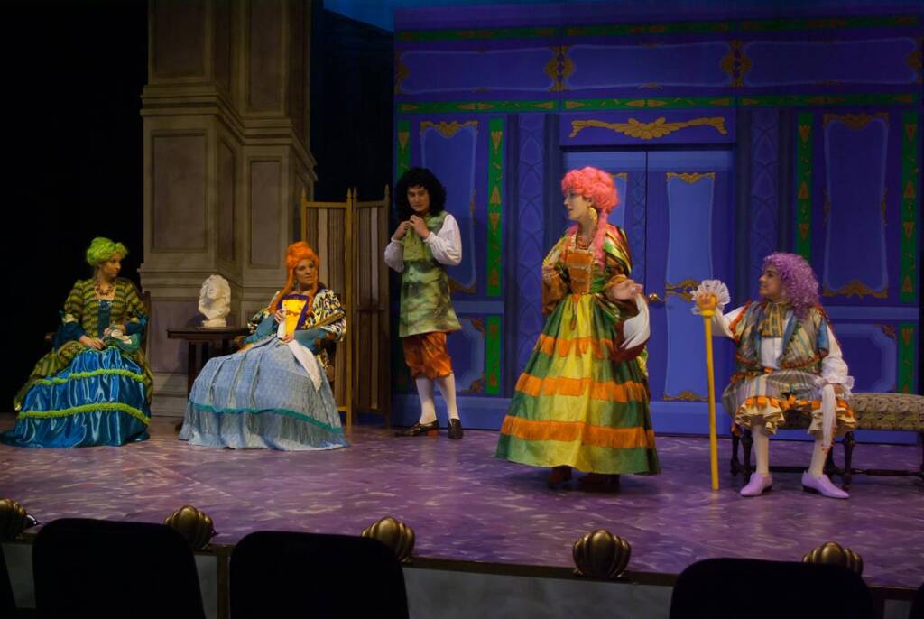 Five actors in colorful, elaborate costumes perform a stage play. They are dressed in ornate, historical outfits, standing in front of a vibrant set with purple and blue decor. One actor stands, gesturing dramatically with a cane.
