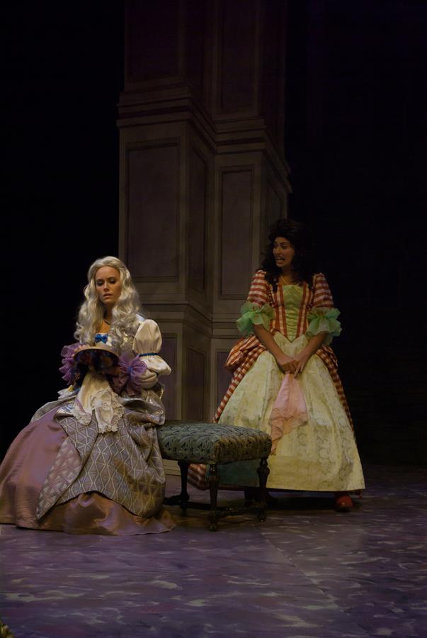 Two women in historical costumes are on stage. One with long, white hair sits in a lavish dress, holding a small object. The other, standing, wears a colorful gown with puffed sleeves and watches her. Both appear to be in a dramatic scene.