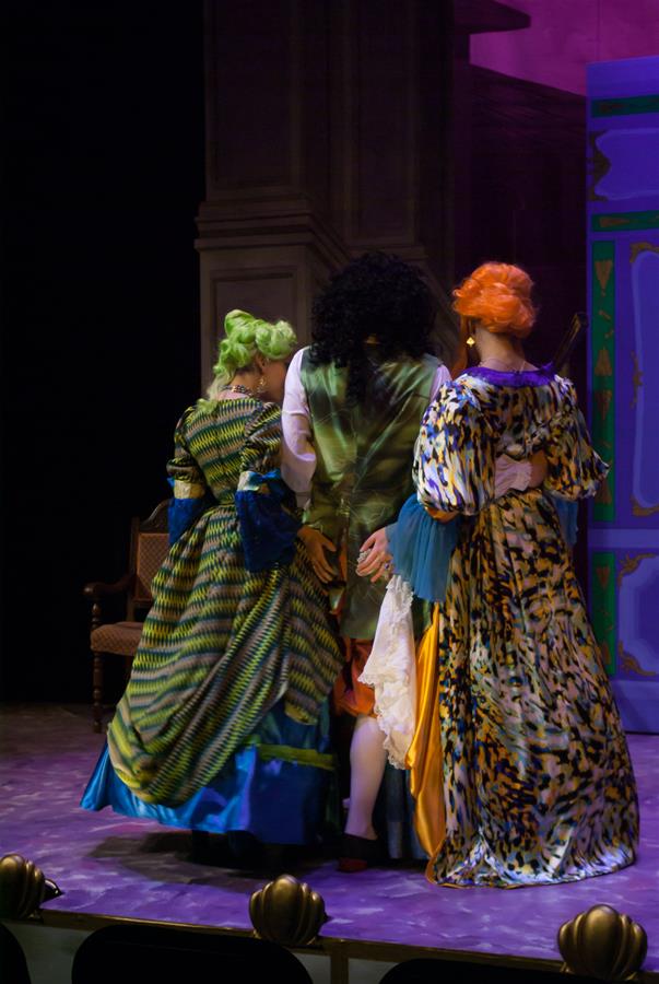 Three actors on stage in colorful, elaborate costumes stand with their backs to the audience. The set behind them features ornate architecture and vibrant colors, enhancing the theatrical atmosphere.