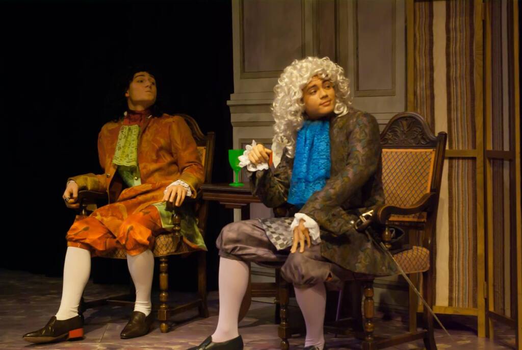 Two actors in elaborate 18th-century costumes sit on stage. One wears a wig and holds a fan, while the other wears a dark wig. The backdrop suggests an ornate interior. They appear engaged in a theatrical performance.