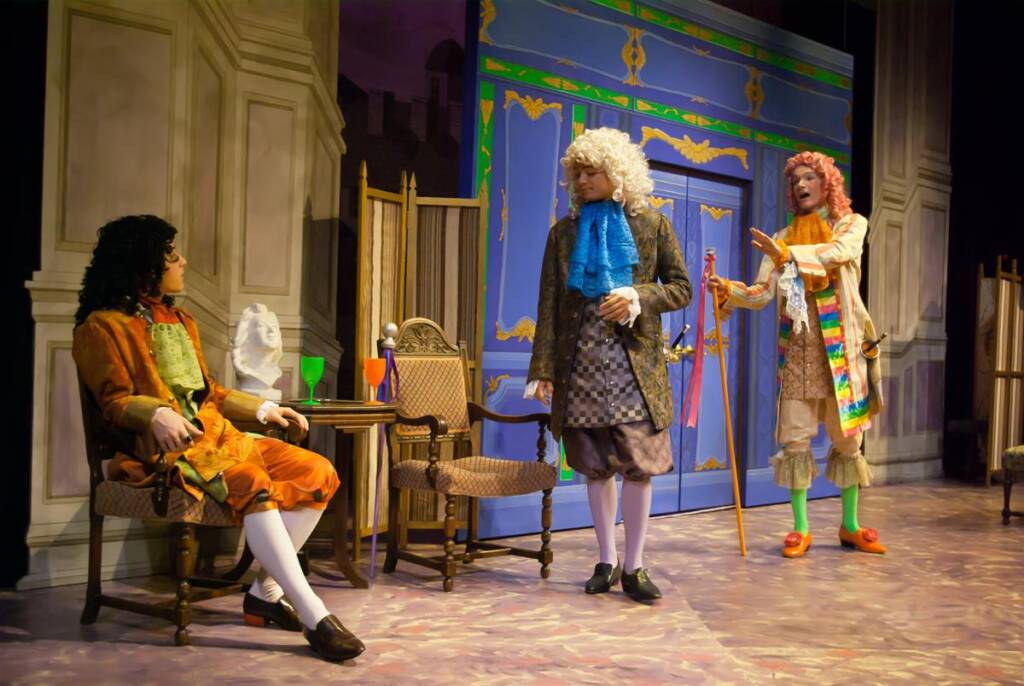 Three actors in 18th-century costumes perform on a stage with ornate set design. One is seated, wearing dark curly hair, another stands with white hair holding a cup, and the third has reddish hair, gesturing expressively with a colorful sash.