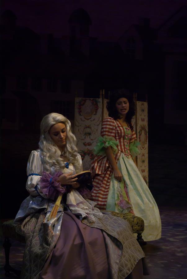 Two women in 18th-century costumes are on stage. One, with long white hair, sits on a bench reading a book. The other, with dark curly hair, stands behind her, gesturing animatedly. A backdrop with a village scene is in the background.