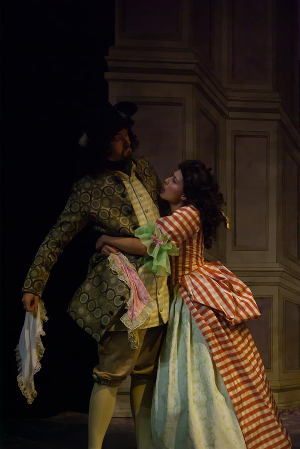 Two actors in period costumes perform on stage. The man wears a patterned suit and holds a handkerchief. The woman wears a red and white checkered dress, embracing him from the side. The background features a tall, decorated wall.