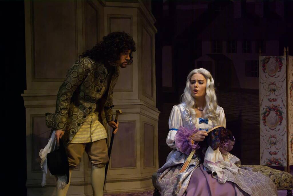 A man in a curly wig and period clothing stands, holding a hat. A woman with long white hair in a historic dress sits, holding a fan. They appear to be in a theatrical play, with vintage-style scenery in the background.