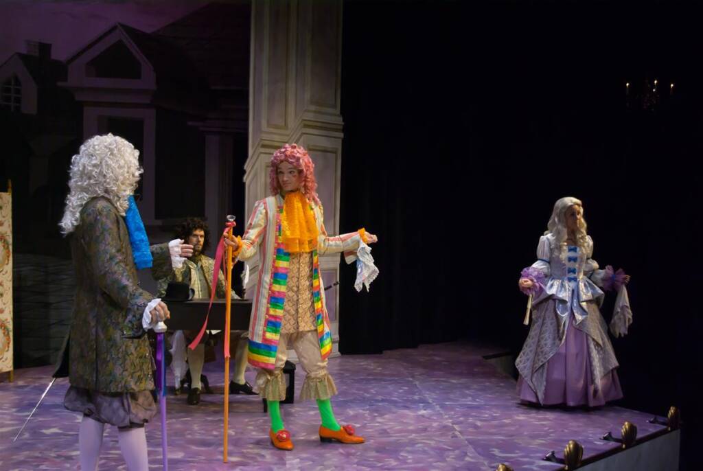 Three actors perform on stage in 18th-century costumes. The central figure wears a colorful outfit with a pink wig, holding a red cane. The other two wear period clothing with wigs. The background features a painted set resembling a classic interior.