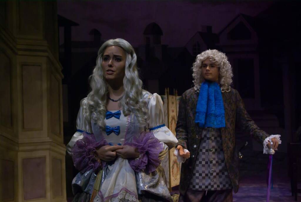 Two actors in period costumes perform on stage. The woman in front wears a white gown with a purple ribbon, while the man behind her wears a dark coat with a blue cravat and a white wig. The backdrop features dimly lit buildings.