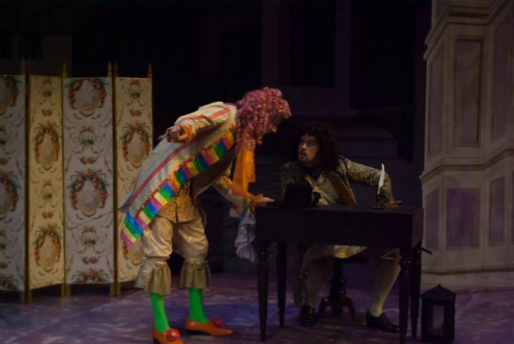Two actors in a theatrical scene. One stands with colorful attire and a pink wig, pointing toward the seated actor in a dark wig and period costume at a small desk. A decorative screen and dim lighting set the stage.