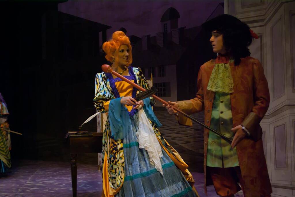 Two actors in ornate historical costumes perform on stage. The woman wears a colorful dress with a blue skirt and orange hair, holding a cane. The man, in a brown coat and wig, gestures with a cane. The backdrop features a street scene.