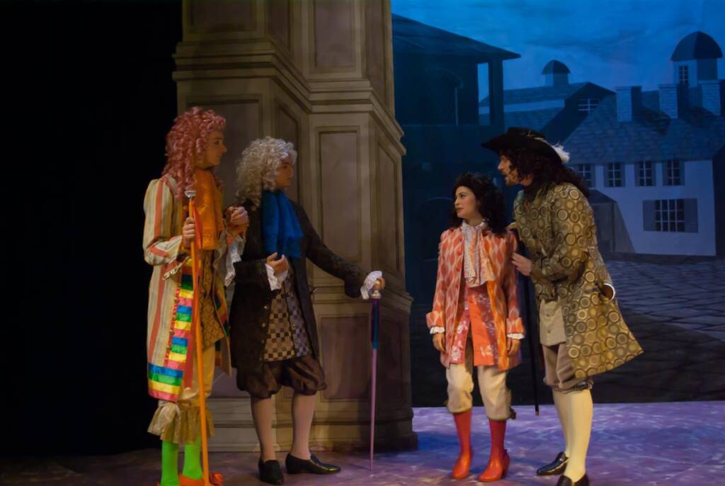 Four actors in 18th-century costumes perform a scene on stage. They wear wigs and elaborate garments, standing near a column with a painted backdrop of a street and buildings. The scene appears dramatic and theatrical.