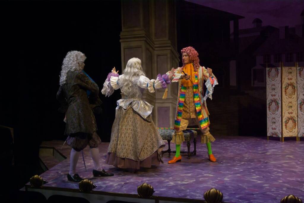 Three actors in elaborate period costumes perform on a theatrical stage. One wears a colorful outfit with green stockings, another in a gray wig and coat. They gesticulate dramatically in front of a column with a painted backdrop.
