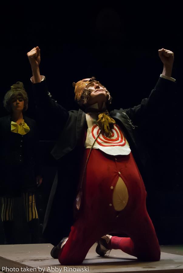 A performer in a colorful, theatrical costume kneels with arms raised triumphantly. The costume features a target design and exaggerated padding. Another performer in a dark outfit stands behind, partially visible. The scene is dimly lit with dramatic effect.