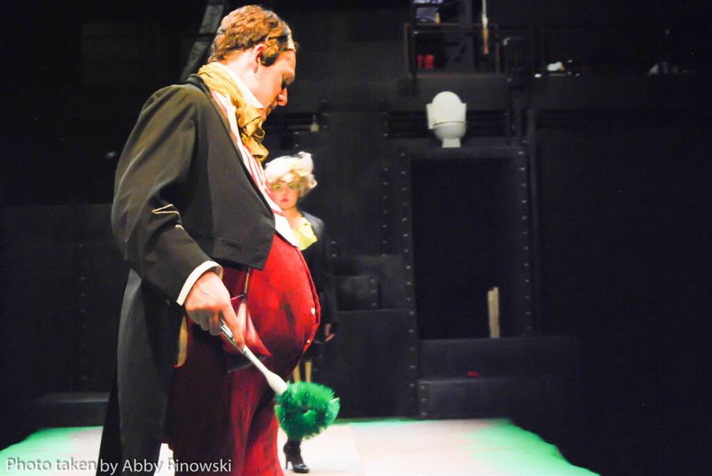 A person in a Victorian-style costume with a long coat and cravat holds a green feather duster, standing on stage. Another person in similar attire is visible in the background. The stage setting includes dark curtains and lighting.