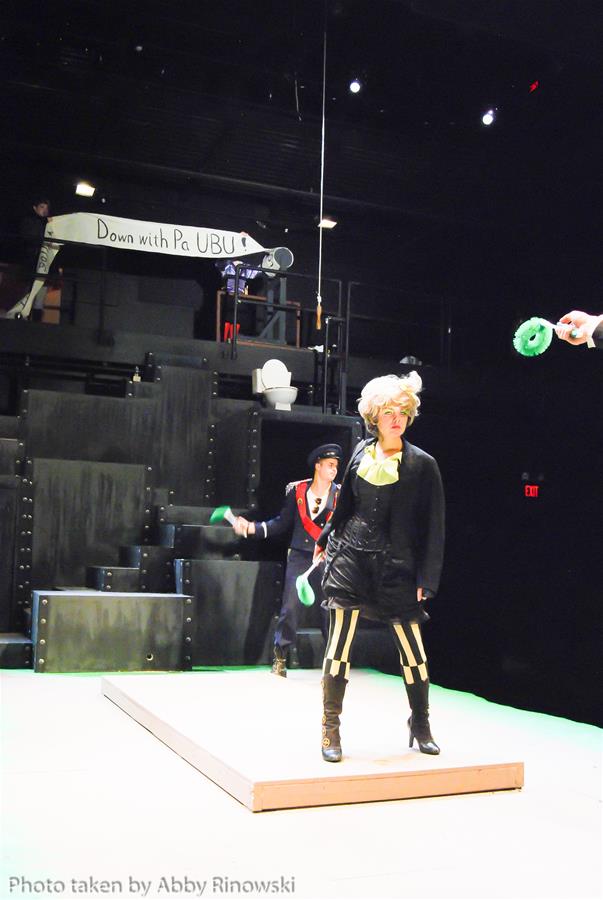 A stage performance with two actors. One in a sailor-like outfit juggles green rings, while the other, with wild hair and striped pants, stands on a platform. A sign reads "Down with Pa UBU" in the background.