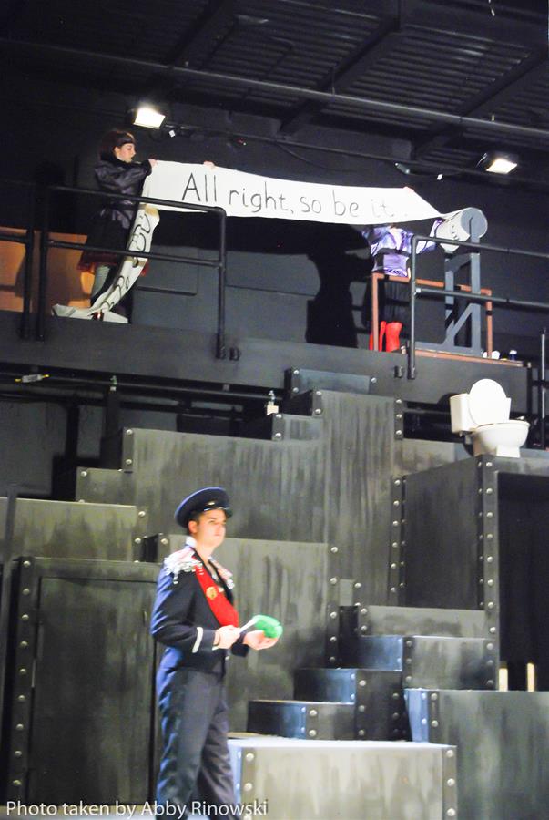 Stage scene from a play features an actor in a military uniform standing below, and two people on a balcony above holding a banner reading "All right, so be it." The set is industrial with dark colors. Photo credit to Abby Rinowski.