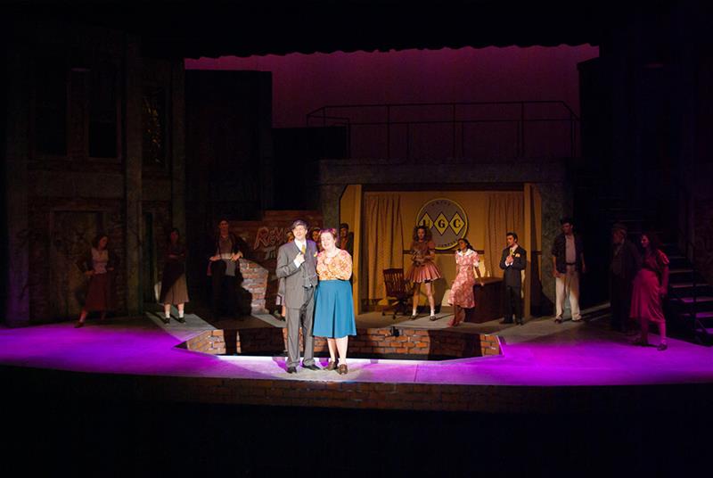 A well-lit stage features a man and a woman in the spotlight, surrounded by other performers in dimmer light. The set includes a backdrop with a yellow sign. The scene exudes a theatrical atmosphere with a mix of colorful outfits.