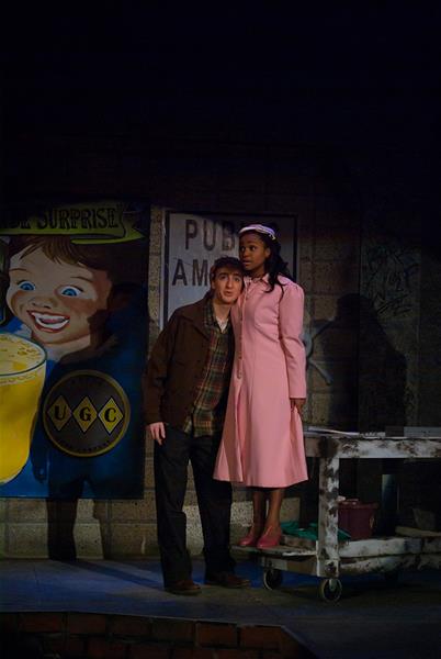 A man in a brown jacket and hat looks concerned while standing next to a woman in a pink coat and hat. They are in a dimly lit room with a vintage sign in the background featuring a smiling child and a drink advertisement.