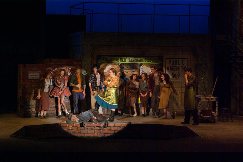 A theater stage with performers in colorful costumes. A person in a bright dress stands over another lying on the ground under a spotlight. Other cast members watch from the background, depicting a lively scene from a play.
