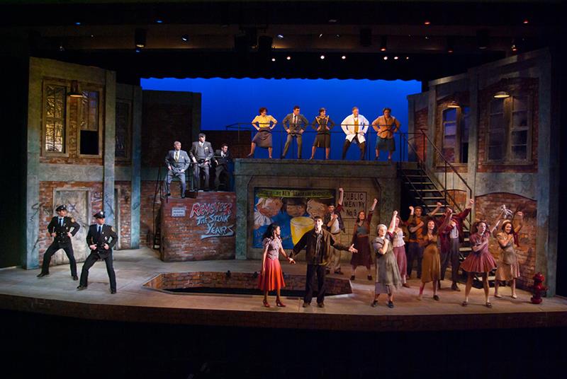 A theatrical scene with actors on stage set in an urban environment. The cast is engaged in a lively performance, with some on a raised platform and others on the main stage. The backdrop features brick buildings and vintage advertisements.