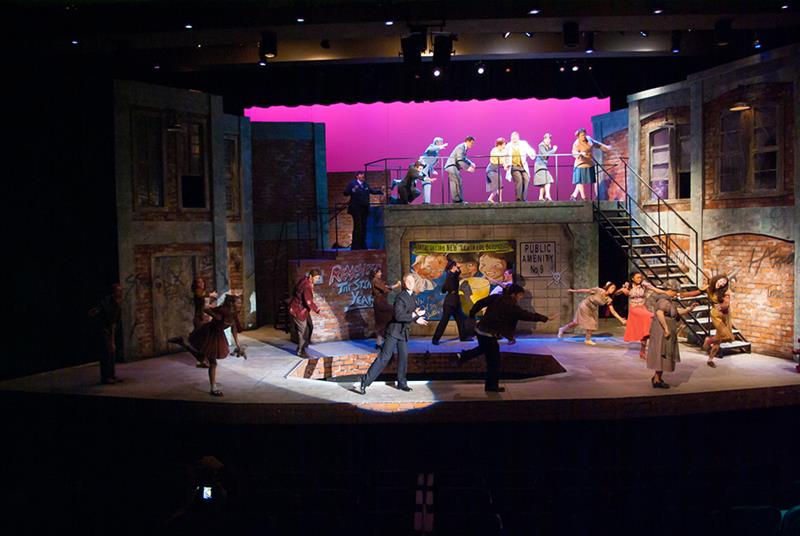 A theater stage shows a lively street scene with actors in 1950s-style outfits. They are engaged in energetic dancing and interactions, depicting a vibrant urban environment. The backdrop features brick buildings and a vibrant pink sky.