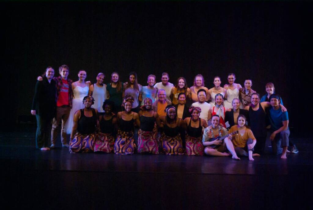 Group photo of a diverse group of performers on stage. The front row features people in colorful, patterned attire kneeling, while others stand behind them. The background is a dark stage, giving the scene a vibrant yet dramatic look.