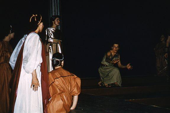 A group of people in ancient Greek-style costumes perform on a dimly lit stage. One person is kneeling with outstretched hands, while others stand and observe. The scene conveys drama and engagement.