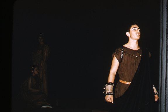 A person in ancient Greek-style attire stands on stage, wearing a dark toga with metallic accents. The background is dimly lit, with two figures partially visible in the shadows, one seated.