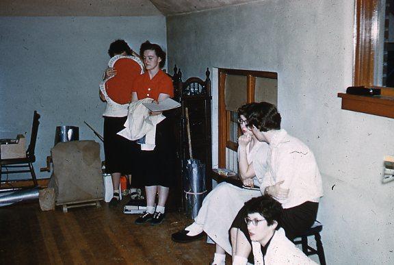 Five women in a room. One hugs a circular red item, another carries clothes. Three sit on a bench; one looks pensive, another whispers, and the third sits on the floor. The room has some clutter, a window, and pale walls.