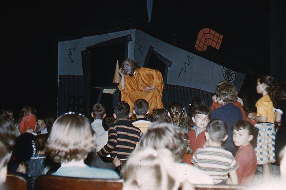 A performer in a yellow costume and gray wig interacts with a group of children in front of a large, cartoonish house backdrop. The children are seated and standing, watching attentively. The scene appears to be a theatrical or puppet show.