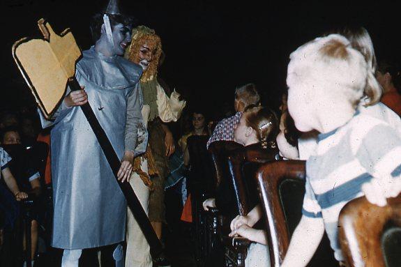 People in costumes resembling characters from "The Wizard of Oz" interact with children seated in wooden chairs. The costumes include a silver figure with an axe and a golden figure with a mane.