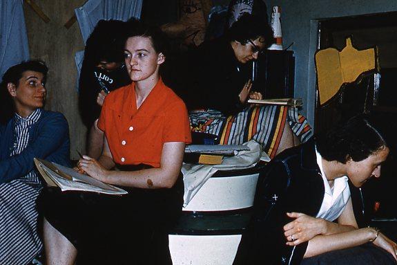 A group of women sitting in a room, focused on drawing or sketching. One woman in the foreground wears a red shirt, while others are behind her, engaged in their work. The setting appears casual, with colorful fabrics in the background.