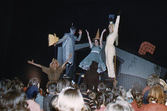 Performers in colorful costumes stand on stage, posing energetically for a lively audience. A backdrop features abstract shapes, and the crowd, mostly children, watch with excitement. The scene is vibrant and theatrical.