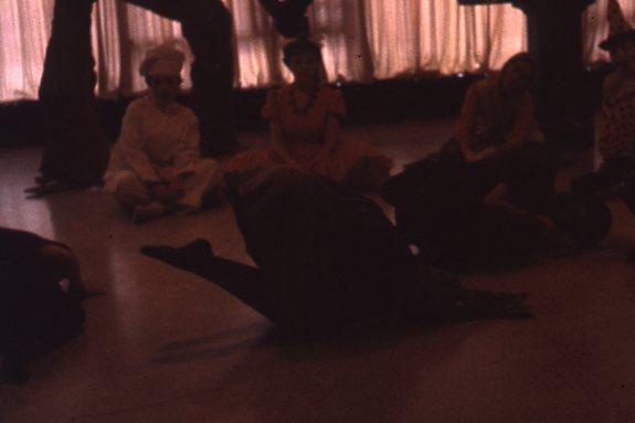 A dimly lit room with people sitting on the floor, some wearing costumes. One person is lying on the ground in the center. The background features draped curtains.