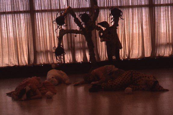 Silhouetted scene of a person standing by a plant with multiple large flowers and four individuals lying on the ground. Light filters through floor-length curtains in the background.