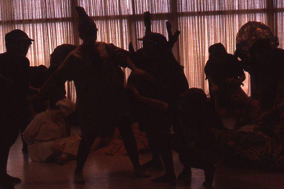 Silhouetted figures of people in elaborate costumes perform in front of a curtain, creating dramatic shadows. The scene suggests a theatrical or dance performance, with various poses and gestures captured.