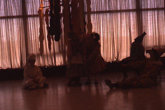 A dimly lit room with large windows has five people in costume. One sits on the floor, two stand, one leans, and another sits with their legs extended. The costumes and lighting create a shadowy, theatrical mood.