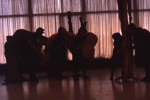 Silhouetted figures in unusual costumes stand or interact in front of a large window with drawn curtains, creating a dramatic play of light and shadow on a smooth, reflective floor.