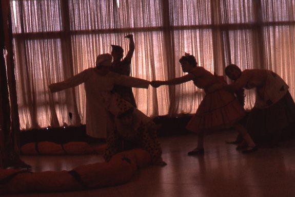 Silhouetted group of five people in costumes perform a dramatic scene near large windows with curtains. A person crouches in the front, while others stand, holding hands and gesturing expressively, in a dimly lit room.