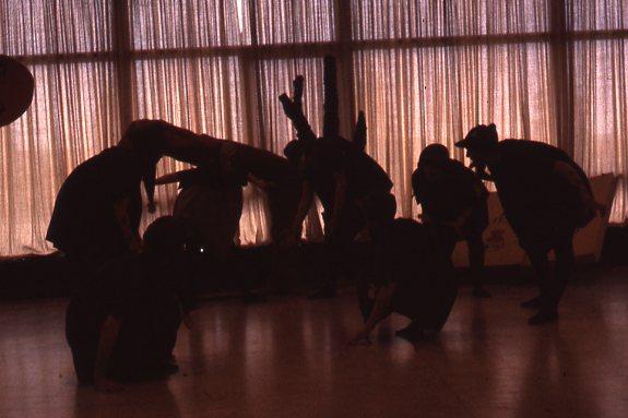Silhouetted figures in dynamic poses gather indoors against a backdrop of large curtains, suggesting a dance or performance. The low light casts shadows, highlighting their movements and creating a dramatic atmosphere.