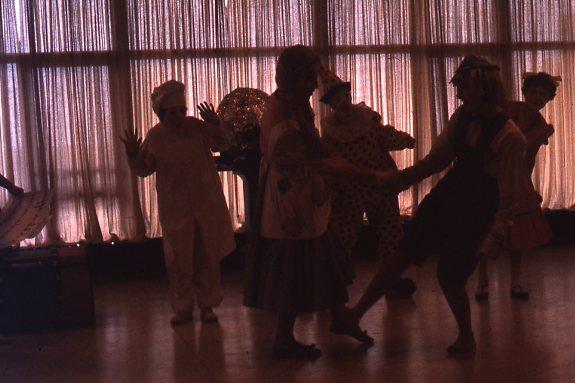 A group of people dressed in costumes dance playfully in a dimly lit room with large windows and sheer curtains, casting soft light.