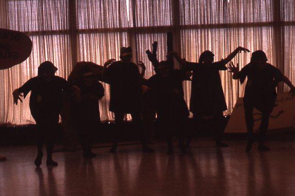Silhouettes of people wearing costumes with outstretched arms, standing in front of large windows with sheer curtains. The lighting creates a dramatic, shadowed effect.