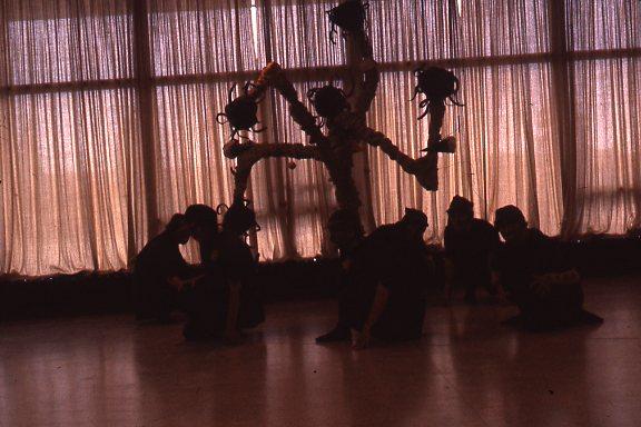 Silhouetted figures kneel on the floor, forming a circle around a central figure with outstretched limbs and abstract structures on their back, against a backdrop of sheer, softly lit curtains.