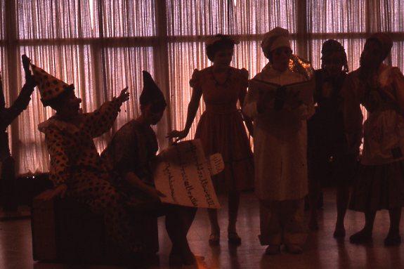 Silhouetted group of people in costume standing indoors. One dressed as a clown sits, holding paper, while others stand nearby. Another person is dressed as a chef, holding a book. Light filters through large windows in the background.