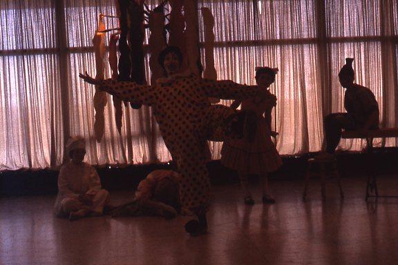 A person dressed as a clown in a polka dot costume balances on one leg in front of a curtain. Children in costumes sit and stand nearby. The room has a dimly lit, playful atmosphere.