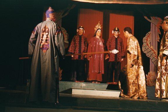 A theatrical performance with actors in elaborate costumes on stage. One actor holds a staff, while others stand in ornate robes and headdresses. The backdrop features a red curtain and decorative elements, suggesting a historical or fantasy setting.