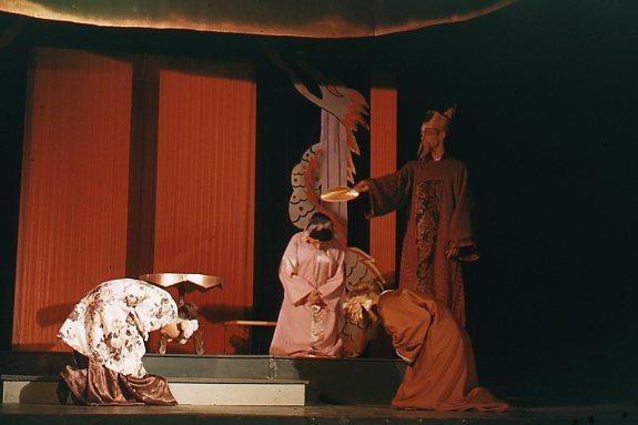 A theatrical scene with four performers in traditional costumes on stage. One stands wearing a robe and hat, holding a platter. The other three are bowing with detailed, colorful attire against a dimly lit, ornately decorated backdrop.