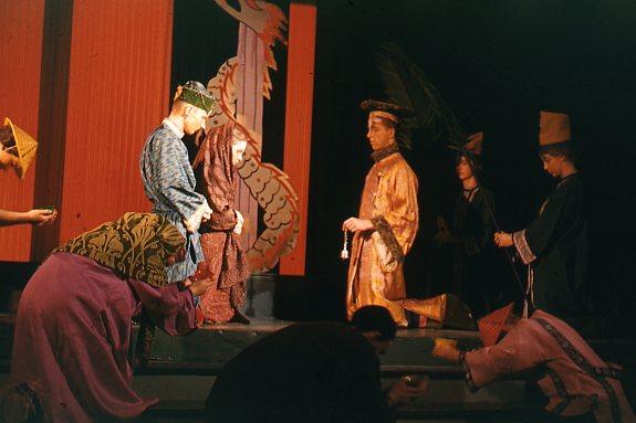 A stage performance featuring actors in traditional Asian-inspired costumes. Some actors kneel while others stand, set against a backdrop with a dragon motif. The scene is dimly lit, evoking a historical or ceremonial atmosphere.