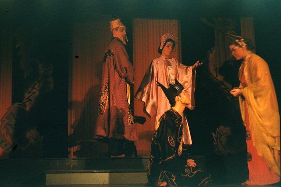 A theatrical scene with four actors in traditional Chinese costumes, featuring intricate robes and headpieces. One actor kneels while another places a crown on their head. The background suggests a richly decorated set.