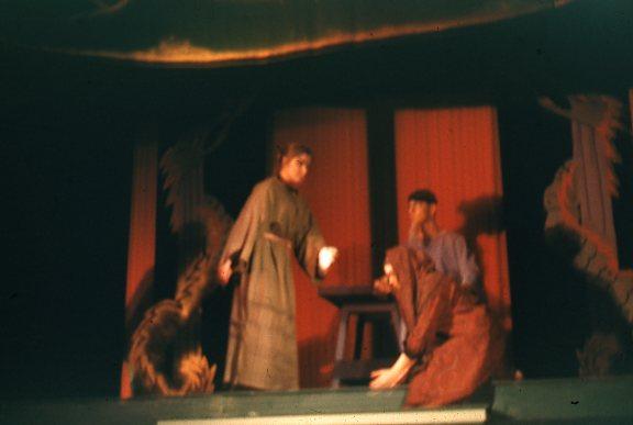 Blurry photo of two people in long robes on a stage with a red and black backdrop. One person stands holding a cup, while the other is seated. Decorative elements suggest an Asian-inspired set design.