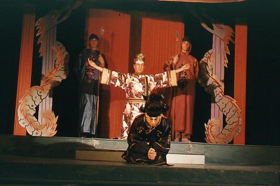 A theatrical scene with four actors in ornate costumes. One performer stands center with outstretched arms, flanked by two others in the background. The fourth actor kneels in the foreground. The setting includes elaborate red and gold drapery.