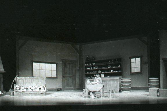 A theatrical stage set depicting a rustic tavern interior. Features include wooden barrels, a table with chairs, a shelf with bottles, and a large washtub. Dim lighting creates a vintage, old-world atmosphere.